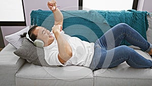 Joyful young chinese man lying on the sofa, confidently listening and dancing to music at home, immersed in the audio world with