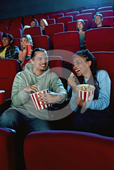 Joyful young couple laughing, having popcorn while watching movie together, sitting in cinema auditorium