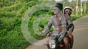 Joyful young couple traveling by motorbike. Two cheerful lovers ride motorcycle on jungle road. Girlfriend smiles, hugs