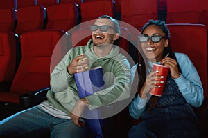 Joyful young couple wearing 3d glasses smiling, while watching a movie together in cinema auditorium, eating popcorn and