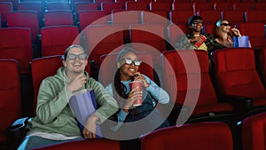 Joyful young couple wearing 3d glasses watching a movie together in cinema auditorium, eating popcorn and drinking soda