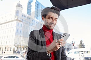 Joyful young elegant dark haired bearded guy holding mobile phone and smiling gladly while checking his social networks, standing