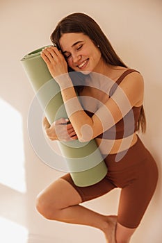 Joyful young european lady clings to yoga mat with her eyes closed while standing near light wall.