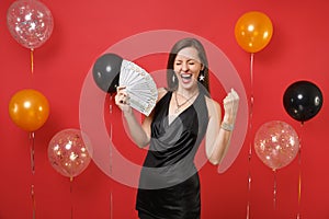 Joyful young girl in black dress screaming, doing winner gesture holding bundle lots of dollars cash money on bright red