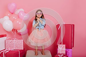 Joyful young girl in blue shirt, tulle skirt holding mask on face suround colorful giftboxes on pink background. Lovely
