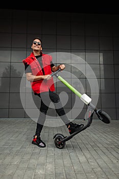 Joyful young hipster man with haircut in stylish denim red-black clothes in sunglasses poses with a modern electric scooter