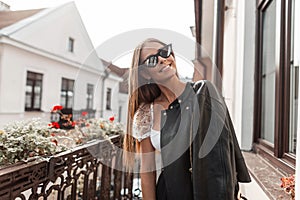 Joyful young hipster woman with a beautiful smile in fashionable sunglasses in a black jacket enjoys relaxation standing on a
