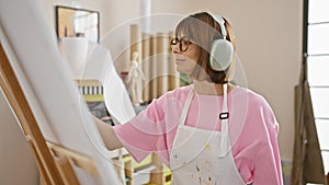 Joyful young hispanic woman artist, swapping paintbrush for headphones, dancing while drawing and listening to upbeat music,