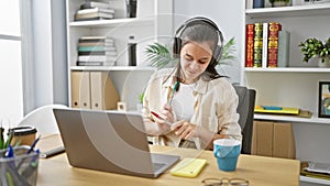 Joyful young hispanic woman worker having fun, dancing to music while working on her laptop at the office, exuding confidence