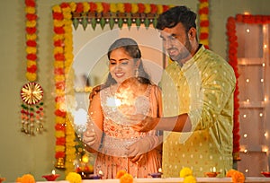 Joyful young indian couples playing with sparklers or firecrackers during diwali festival celebration at home - concept