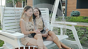 Joyful young ladies friends swinging having fun on swing outside in city on summer day