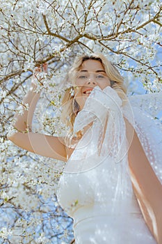 Joyful young lady in wedding attire with big bow on shoulder standing under white sakura and holding branch of tree.