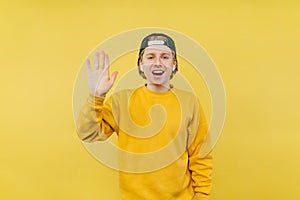 Joyful young man in a colored sweatshirt and cap shows a gesture of congratulations with his palm to the camera and looks at the