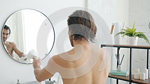 Joyful young man dancing in bathroom with towel looking at mirror reflection