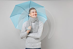 Joyful young man in gray sweater, scarf looking aside, holding blue umbrella isolated on grey background. Healthy