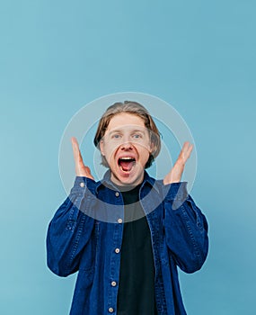 Joyful young man in a shirt stands on a blue background with arms raised and shouts with happiness looking at the camera, isolated