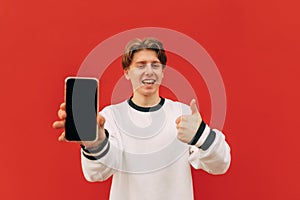 Joyful young man showing thumb up and smartphone screen with camera on red wall background, smiling and looking at camera