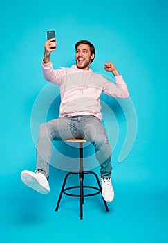 joyful young man sitting on chair and holding mobile phone isolated over light blue, celebrating financial success