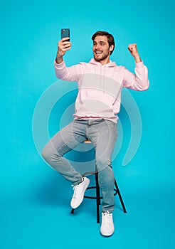 joyful young man sitting on chair and holding mobile phone isolated over light blue, celebrating financial success