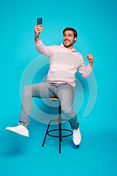 joyful young man sitting on chair and holding mobile phone isolated over light blue, celebrating financial success