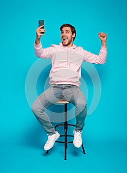 joyful young man sitting on chair and holding mobile phone isolated over light blue, celebrating financial success