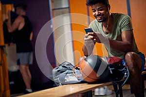 Joyful young man sitting in gym dressing room, looking at smartphone, preparing for workout