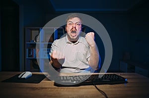 Joyful young man sitting at night at the computer at work and emotionally happy with a smile on his face