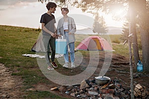 Joyful young man and woman standing at camping stop with portative fridge and laughing.