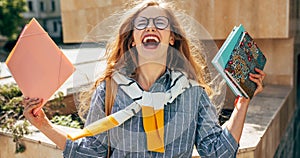 A joyful young student woman standing outdoors next to the college campus with hands raised with books and folders celebrate her