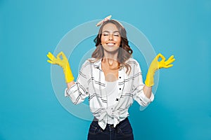 Joyful young woman 20s showing meditation sign with fingers while wearing yellow rubber gloves for hands protection during