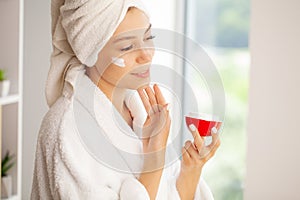 Joyful young woman applying moisturizing cream on face