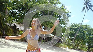 Joyful young woman in bikini running along the exotic white sandy beach on sunny summer day and splashing water. slow