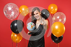 Joyful young woman in black dress celebrating holding world globe, pointing finger on camera on red background air