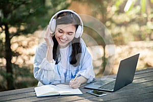 A joyful young woman enjoys her music with headphones, writing in a notebook beside her laptop