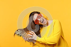 Joyful young woman in funny glasses with volant hair holding fresh ripe pineapple fruit isolated on yellow orange wall