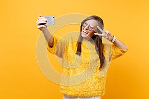 Joyful young woman in fur sweater heart glasses showing victory sign doing taking selfie shot on mobile phone isolated