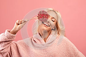 Joyful young woman holding candy on stick, covering one eye with lollipop and smiling, having fun over pink background