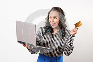 A joyful young woman holding a plastic Bank card and a laptop. Studio photo on white background