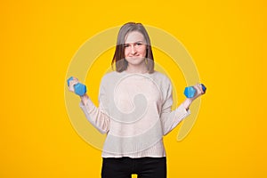 A joyful young woman is looking at the camera smiling and is working out with two dumbbells