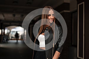 Joyful young woman with natural make-up with brown hair in a stylish leather black jacket in a trendy top stands in the city near