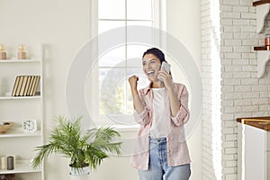 Joyful young woman sincerely rejoices hearing good news while talking on mobile phone at home.