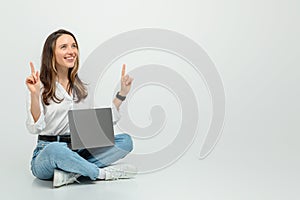 Joyful young woman sitting cross-legged on the floor, holding a laptop with one hand