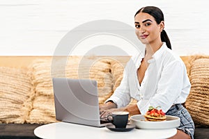 Joyful young woman sitting at table with laptop near delicious pancakes with berries