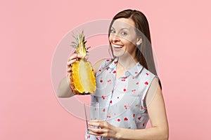 Joyful young woman in summer clothes holding half of fresh ripe pineapple fruit, glass cup  on pink pastel wall