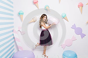 Joyful young woman in tulle skirt having fun with big macaron on white background among sweets. Pastel colors, ice cream