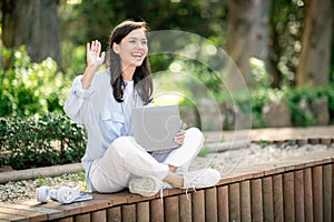 Joyful young woman waving while seated on a park bench, holding a laptop in her lap