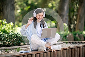 Joyful young woman wearing headphones waving at her laptop screen during a video call