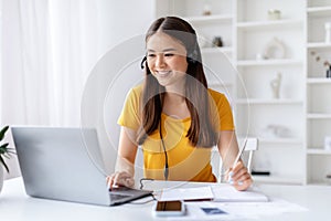 Joyful young woman wearing headset multitasking with pen and laptop at home