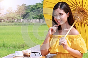 Joyful young women wear yellow dresses holding yellow umbrella sitting relax with camera, cup of coffee and cakes looking at