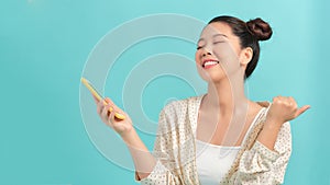 Joyous asian woman with big smile chatting using cell phone and showing thumb up isolated over turquoise background